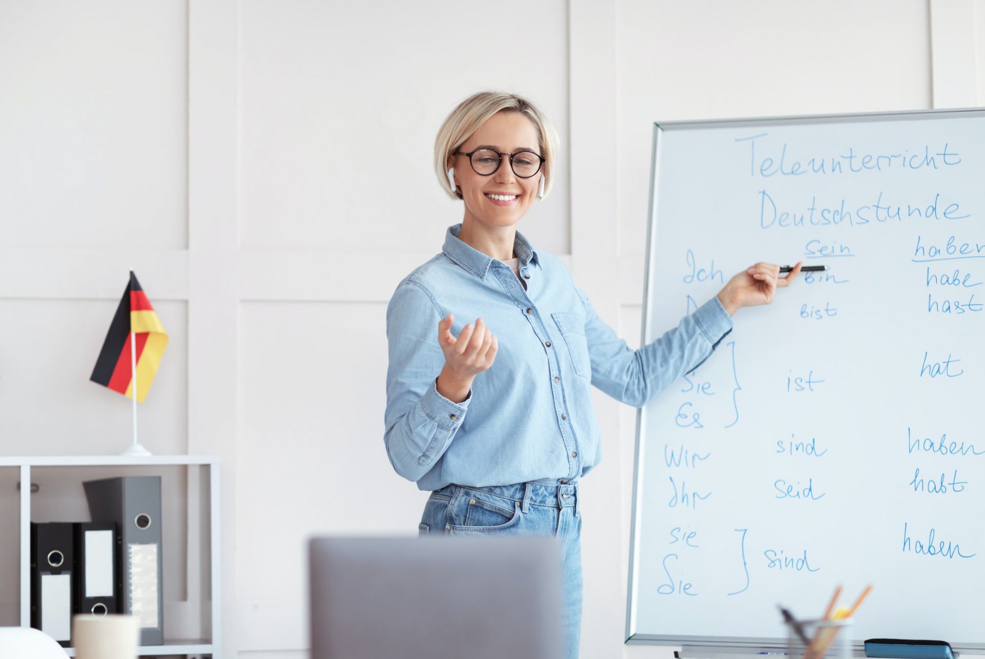 Friendly young female teacher giving German lesson online on laptop, pointing at blackboard with